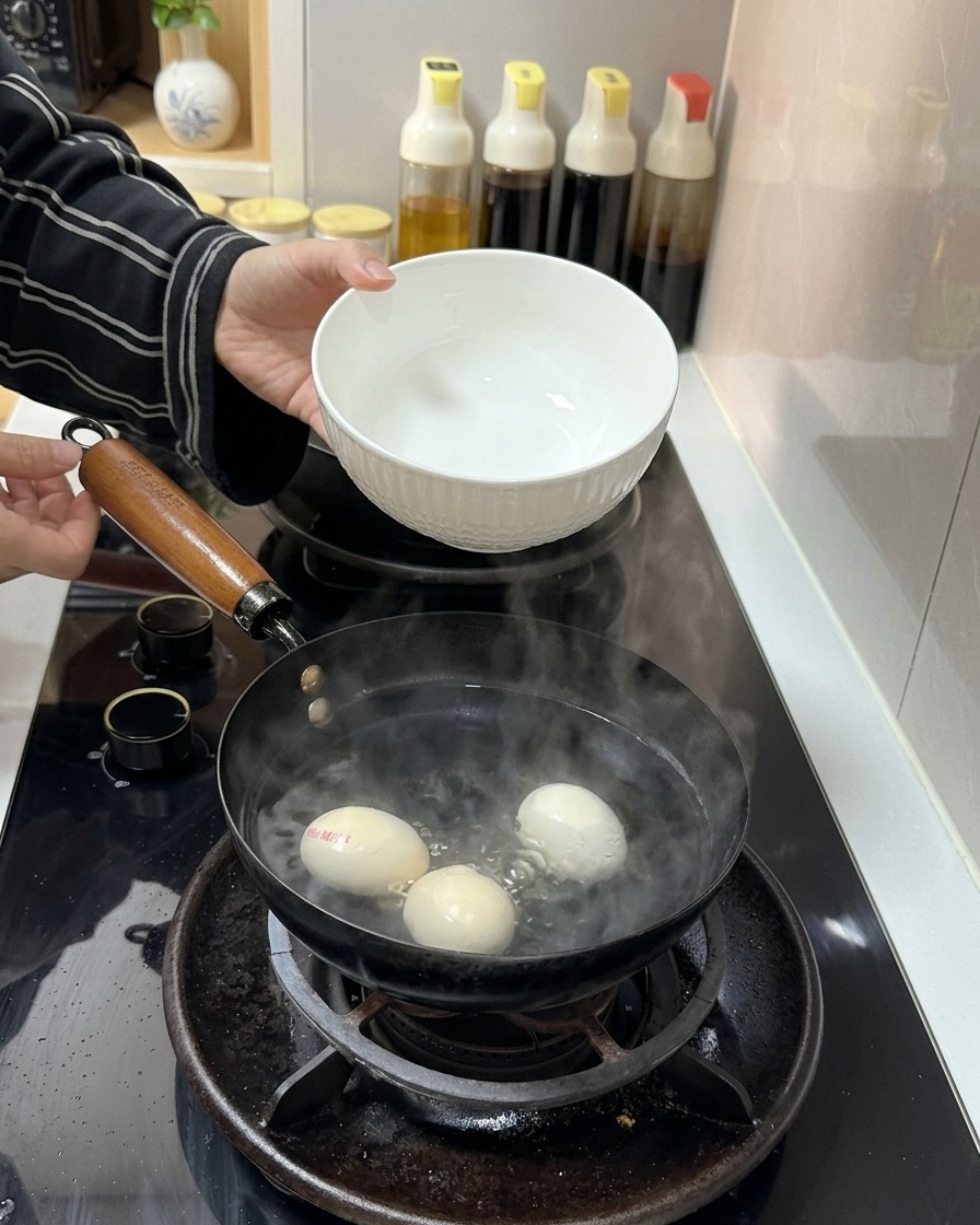 Un bol en céramique blanche versant de leau froide dans une casserole contenant de leau bouillante et trois œufs.