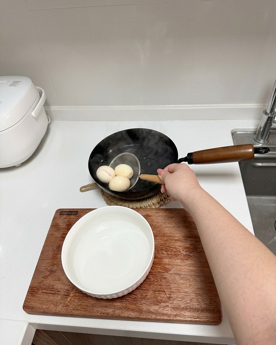 Une main utilisant une petite passoire pour retirer trois œufs de la casserole vers un bol deau froide.