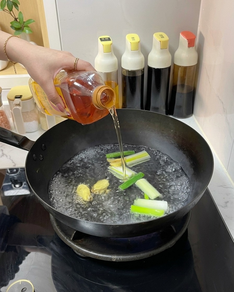 Une personne versant un liquide ambré depuis une bouteille en plastique dans un wok deau bouillante avec du gingembre et des oignons verts.