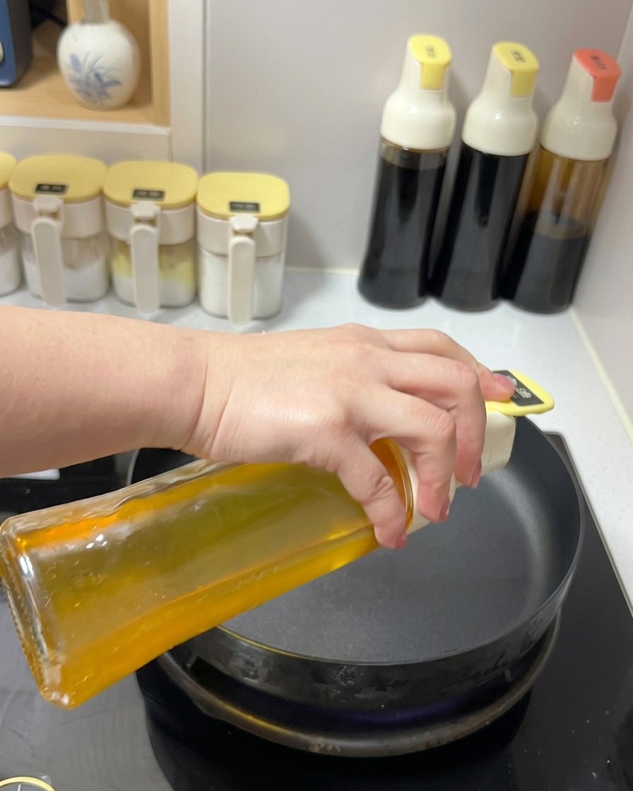 Huile de cuisson transparente versée depuis un flacon en verre dans une poêle noire sur une plaque de cuisson.