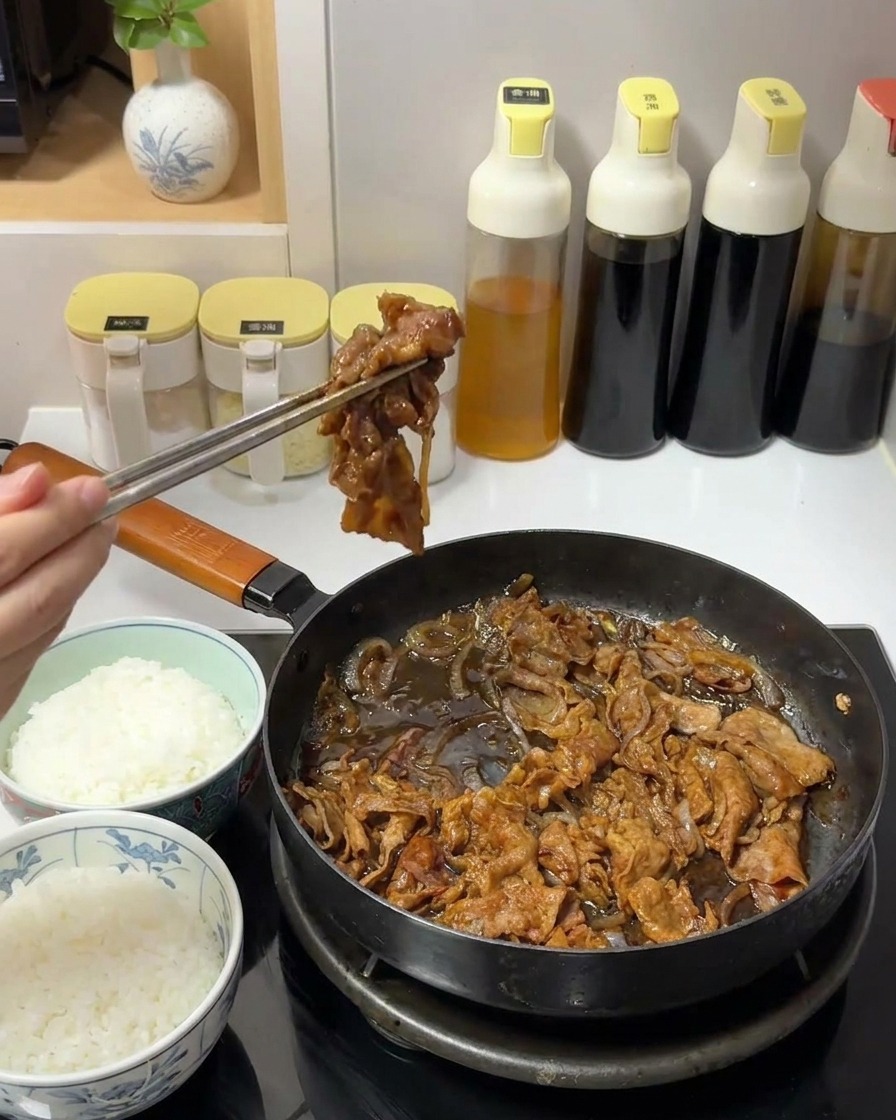 Une main utilisant des baguettes en métal pour prélever une portion de bœuf en sauce et doignons dans une poêle.