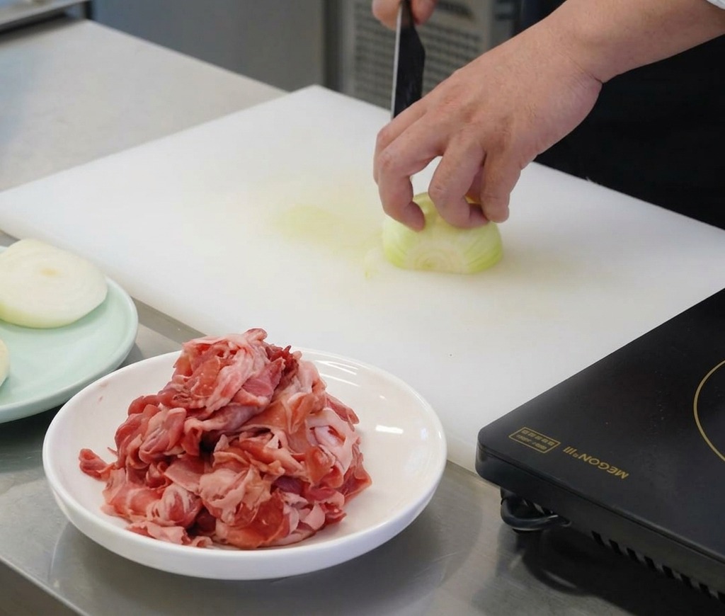 Une main de chef maintenant un oignon blanc sur une planche à découper blanche tout en le coupant en fines lamelles verticales avec un couteau bien aiguisé.