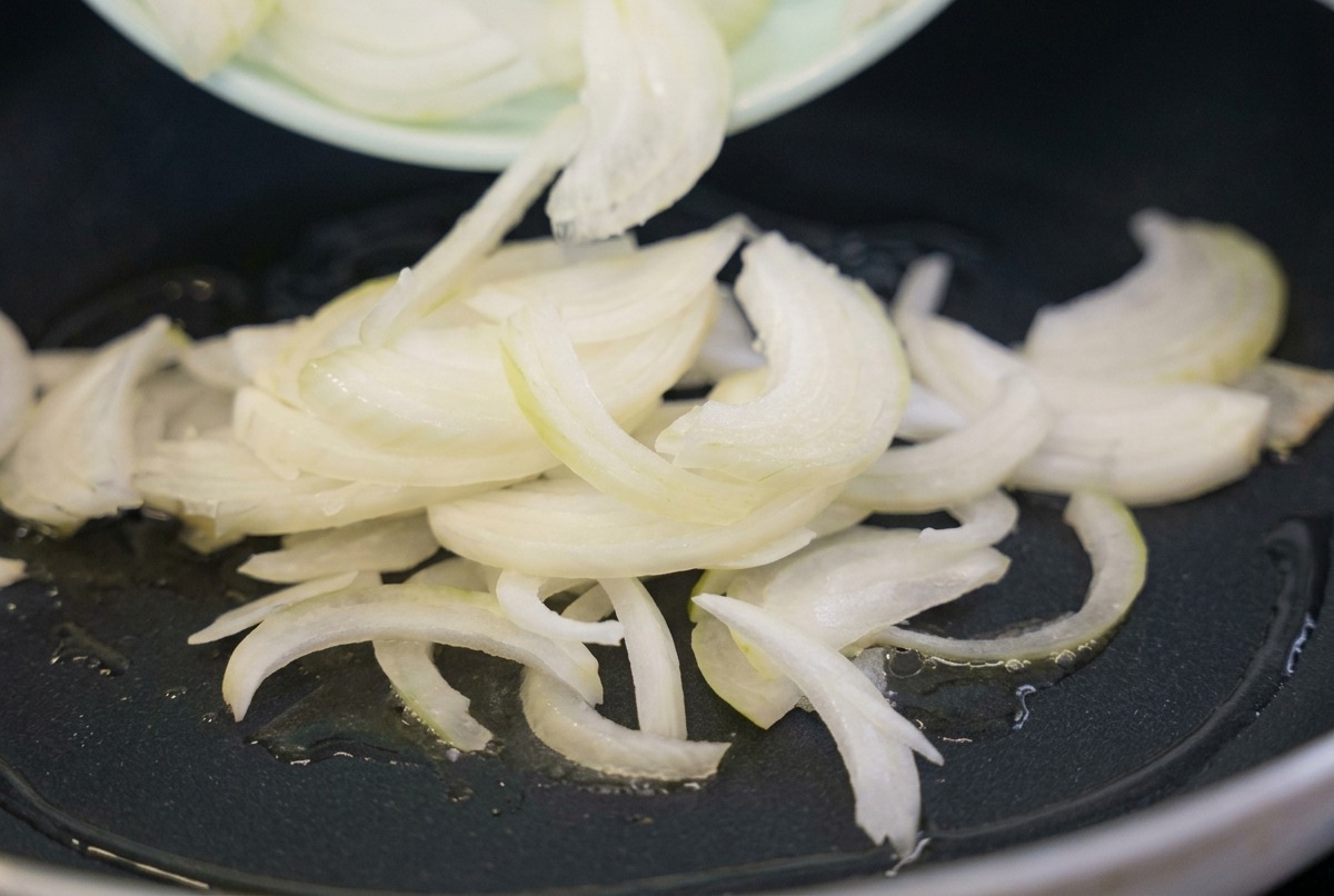 Des lamelles doignon blanc cru versées depuis une assiette vert clair dans une poêle noire contenant de lhuile de cuisson chaude.