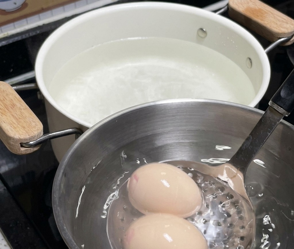 Deux œufs reposant dans un bol en acier inoxydable rempli deau glacée, avec la casserole blanche en arrière-plan.