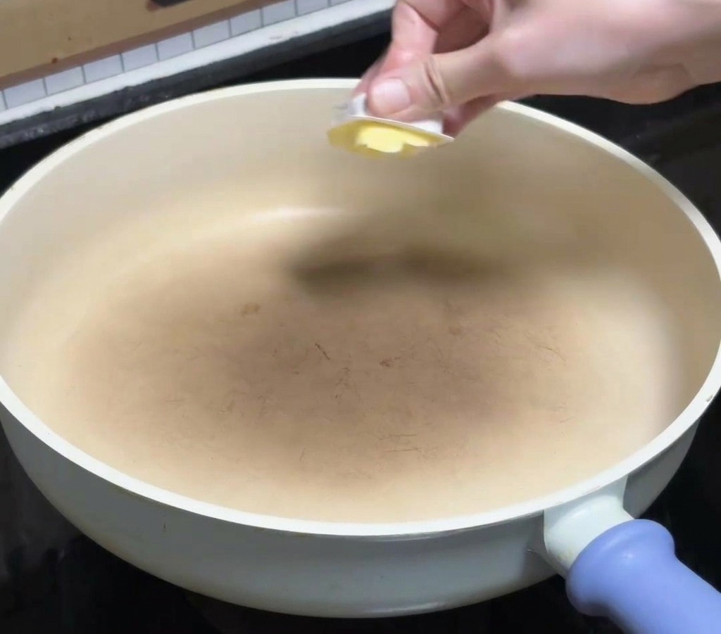 Une main déposant un morceau de beurre jaune dans une poêle en céramique blanche propre sur une plaque de cuisson.