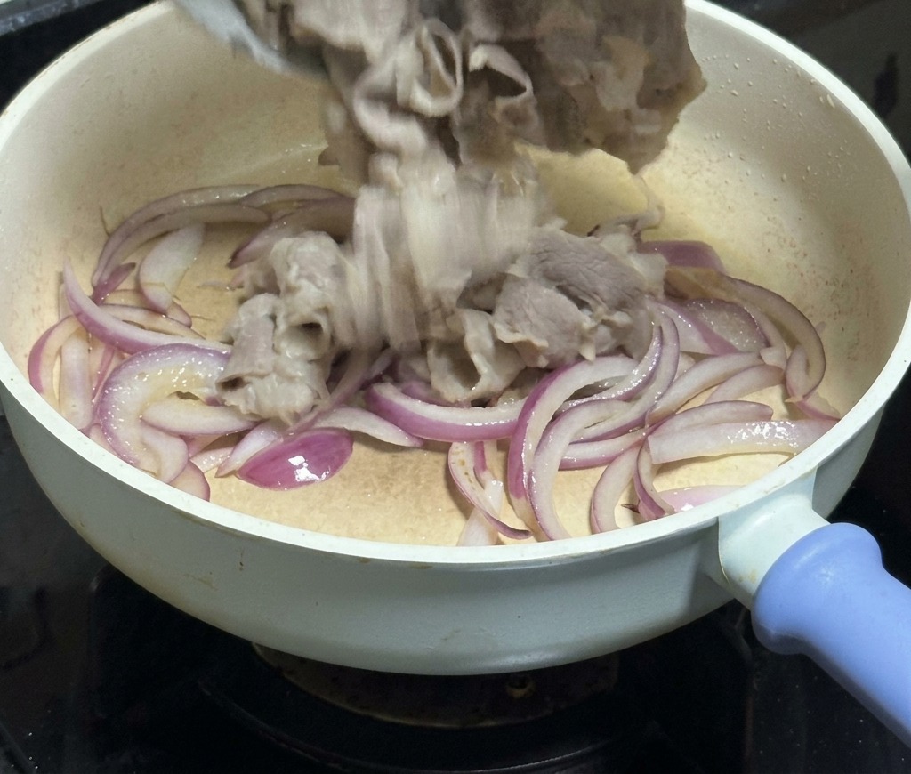 De fines tranches de bœuf versées depuis un bol blanc dans une poêle remplie de lamelles doignon rouge translucides.