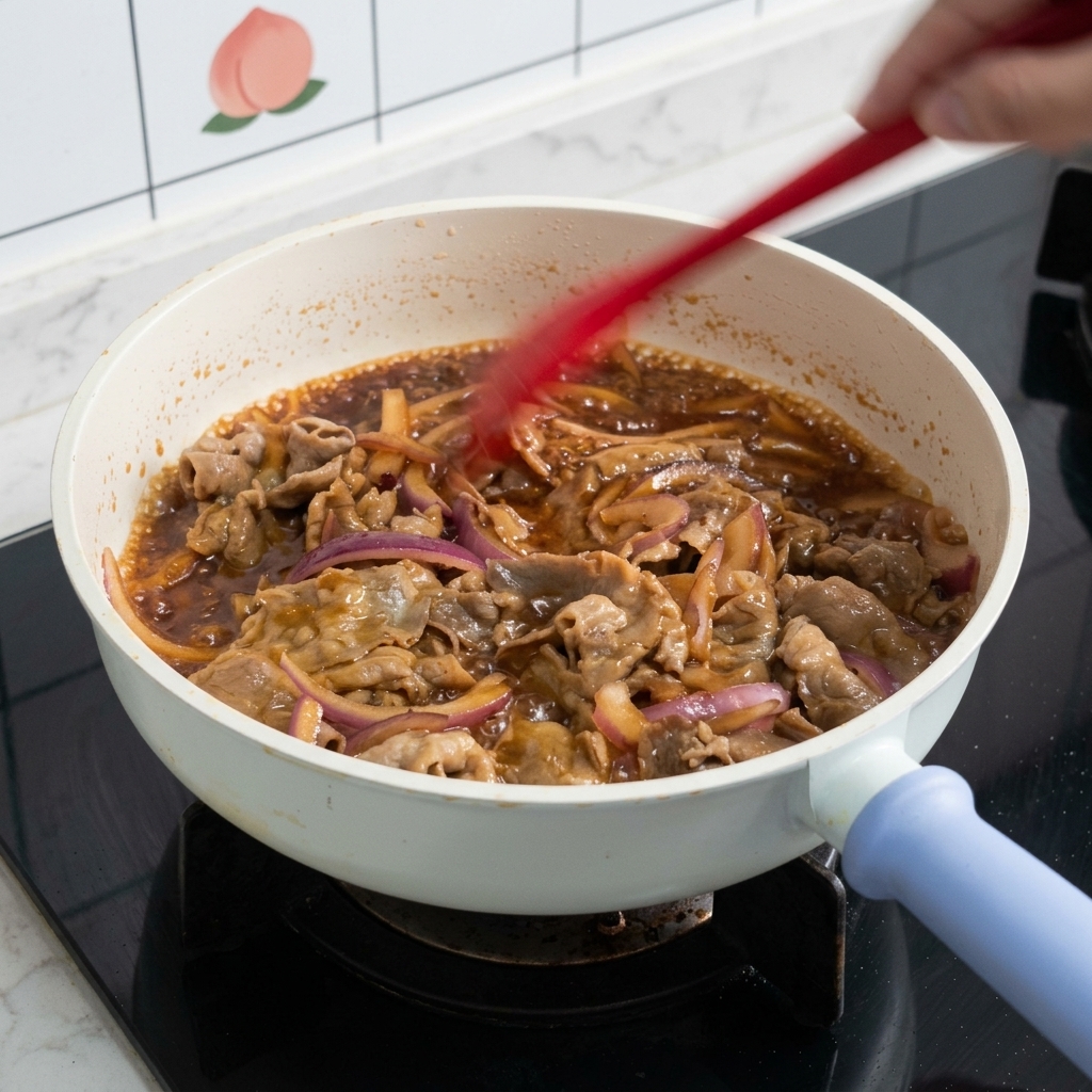 De fines tranches de bœuf et des rondelles doignon rouge mélangées avec une spatule rouge dans une poêle blanche remplie dune sauce teriyaki brune et bouillonnante.