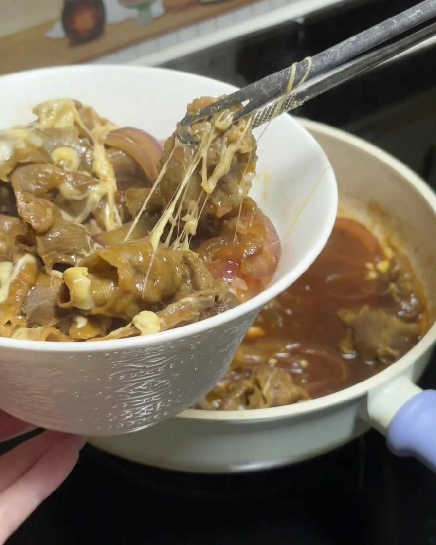Une pince soulevant une portion de bœuf teriyaki au fromage fondant et aux oignons depuis la poêle, laissant filer de longs fils de fromage, pour la déposer sur un bol de riz blanc.