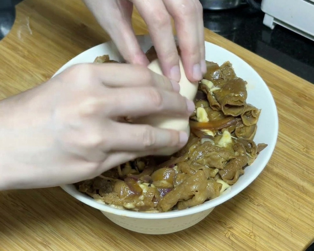 Deux mains cassant délicatement un œuf onsen mollet sur un bol de bœuf teriyaki recouvert de fromage fondu.