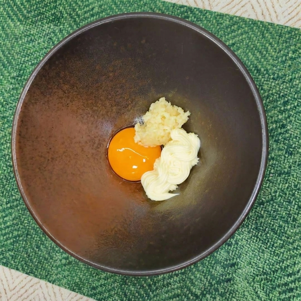 Un bol en céramique contenant un jaune dœuf, une dose de mayonnaise et de lail émincé.