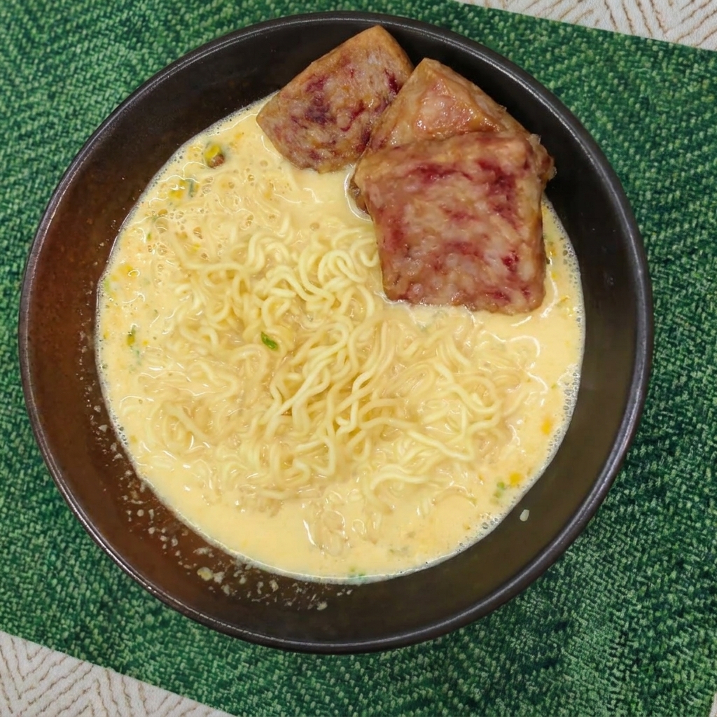 Nouilles dans un bouillon crémeux garnies de trois tranches de spam bien dorées dans un bol sombre.