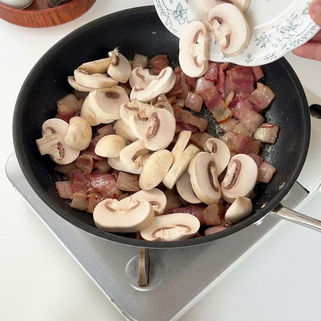 Lamelles de champignons de Paris ajoutées dans une poêle chaude avec du bacon et des oignons.