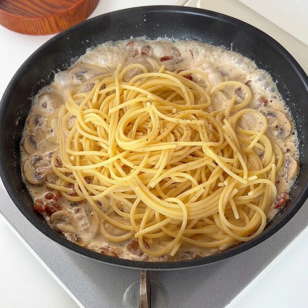 Spaghettis cuits déposés sur la sauce crémeuse dans la poêle et saupoudrés de poivre.