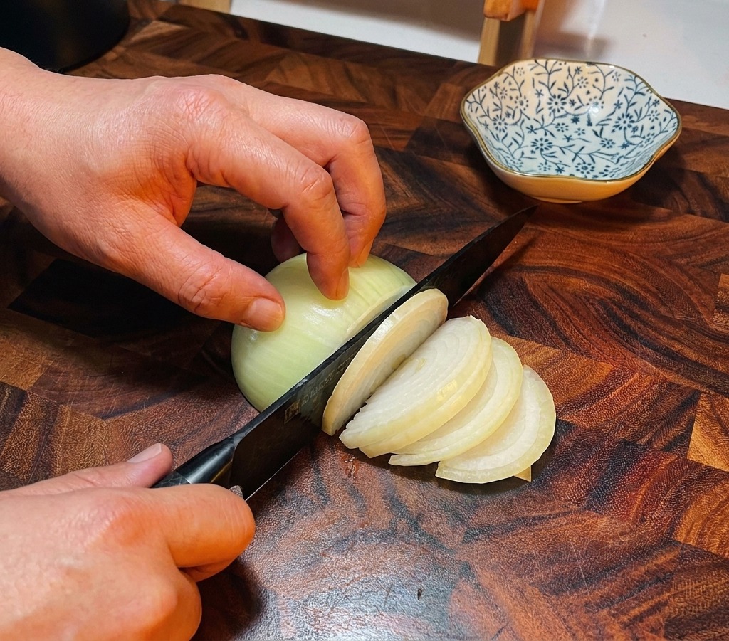 Un couteau de chef éminçant un oignon blanc sur une planche à découper en bois foncé.