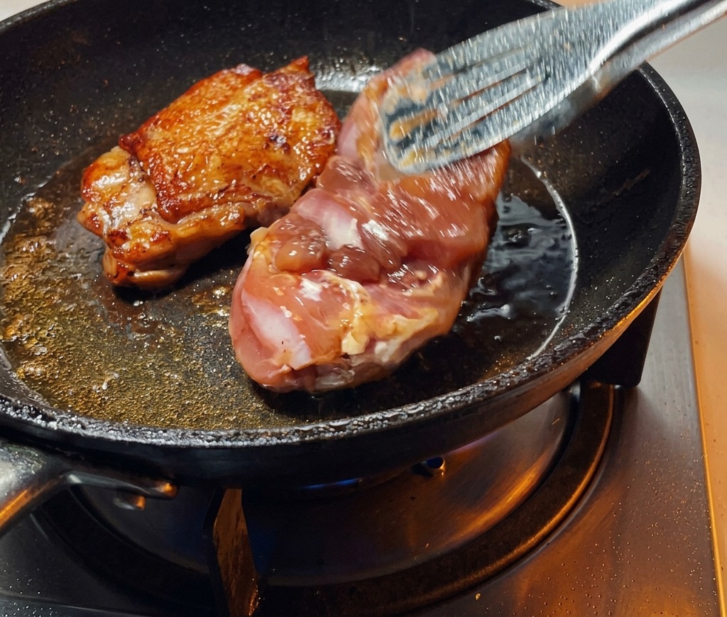 Une pince retournant un haut de cuisse de poulet saisi dans une poêle, révélant une peau parfaitement dorée et croustillante.