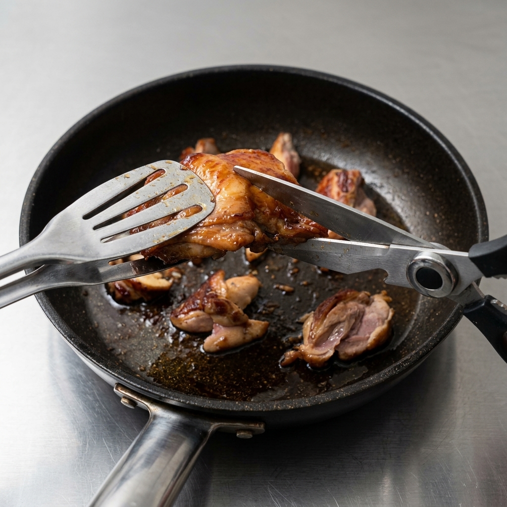 Des ciseaux de cuisine coupant des hauts de cuisse de poulet saisis directement dans la poêle pour former de petits morceaux uniformes.