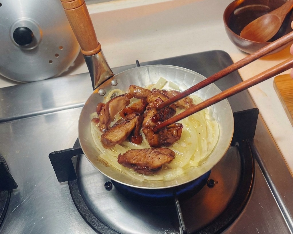 Morceaux de poulet saisis disposés avec des baguettes sur un lit doignons translucides dans une petite poêle.
