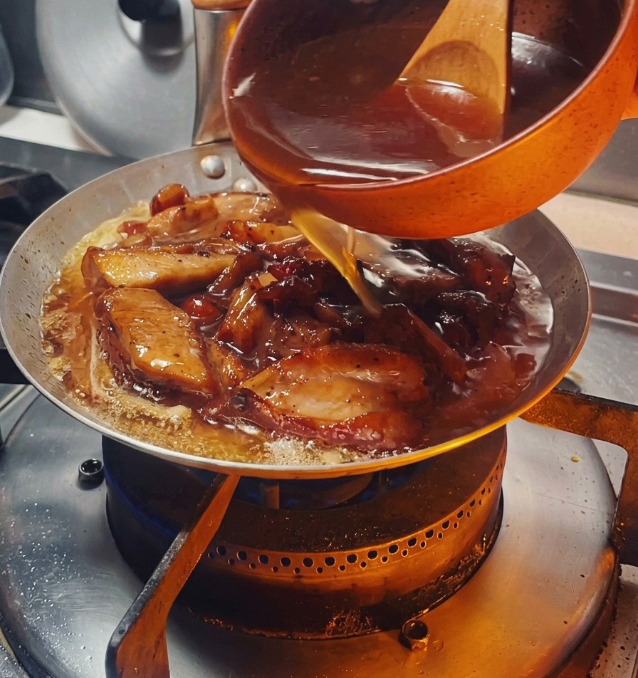 Une sauce sombre et savoureuse versée depuis un petit bol orange dans une poêle remplie de morceaux de poulet dorés et doignons.