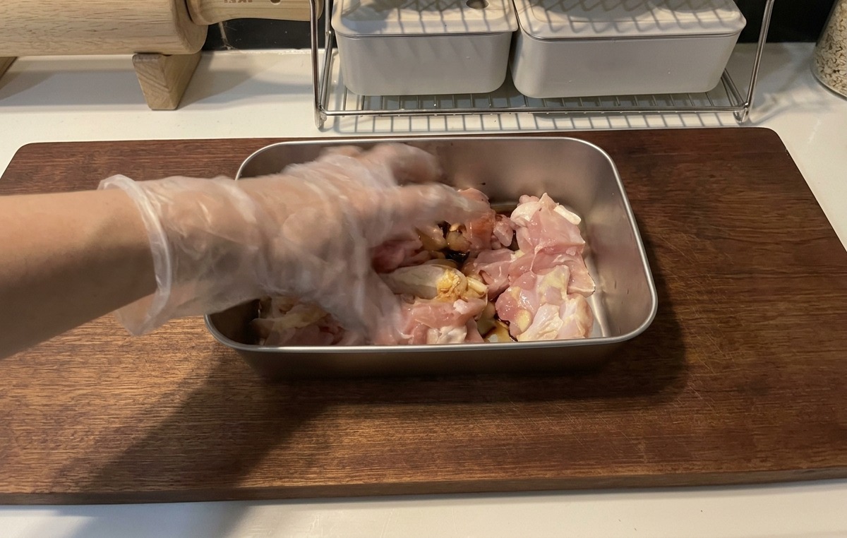 Une main avec un gant plastique mélangeant les morceaux de poulet avec une marinade sombre dans un plat métallique.