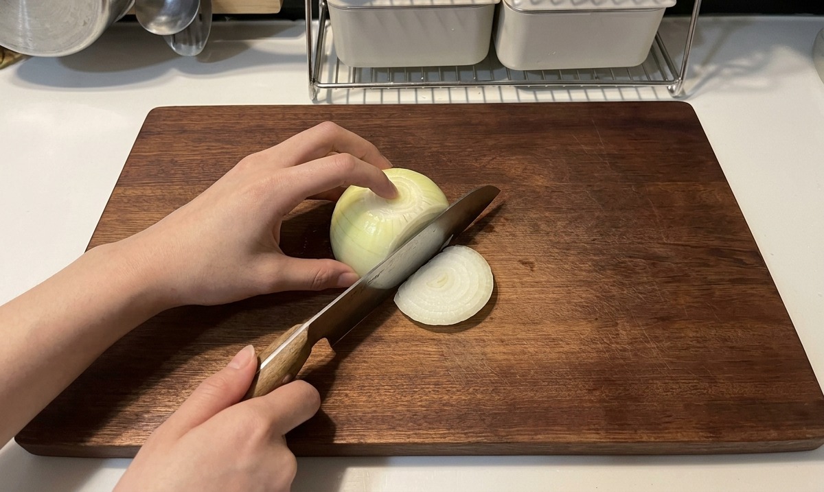 Des mains utilisant un couteau au manche en bois pour émincer un oignon blanc en fines lamelles sur une planche sombre.