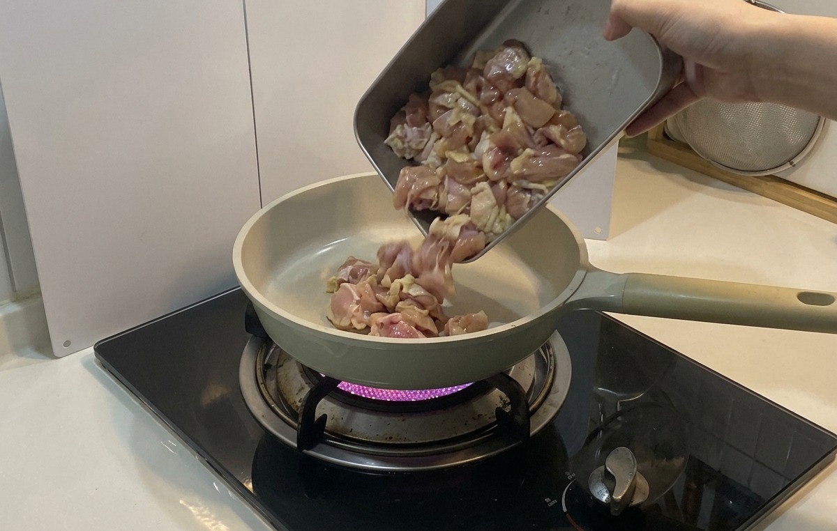 Les morceaux de poulet mariné versés depuis un plat en métal dans une poêle en céramique claire sur un feu de gaz.