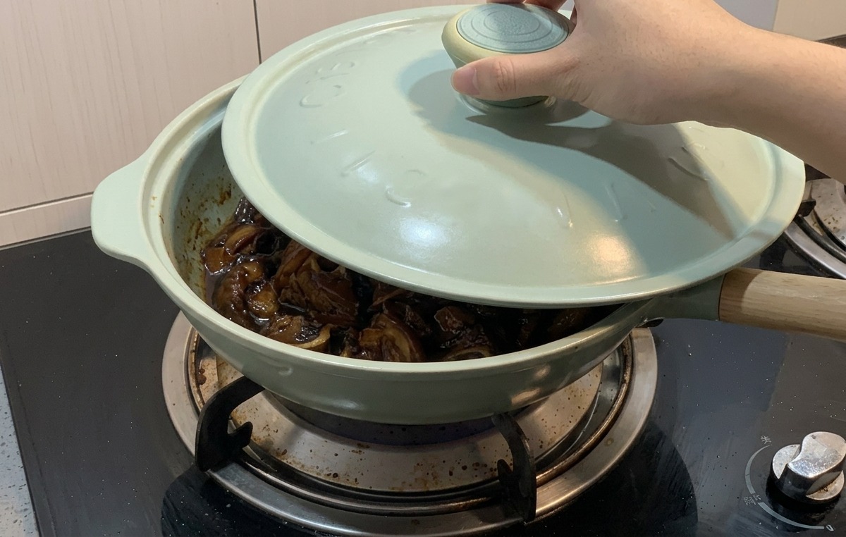 Une main posant un couvercle bleu clair sur une poêle verte où mijotent le poulet et les oignons dans une sauce sombre.