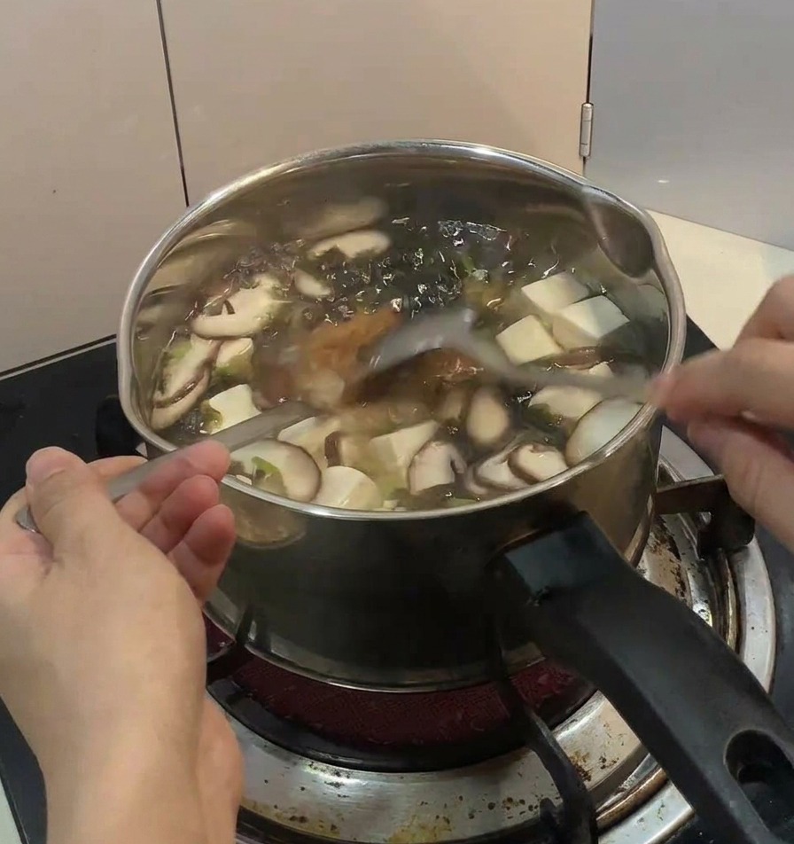 Une main parsemant de la ciboule fraîche et des filaments de nori sur un bol fumant de poulet et dœuf sur table en bois.