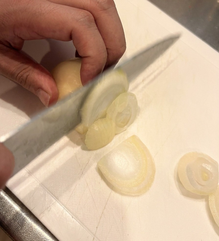 Un couteau aiguisé éminçant un oignon blanc en fines lamelles sur une planche à découper blanche, avec une main maintenant loignon en sécurité.