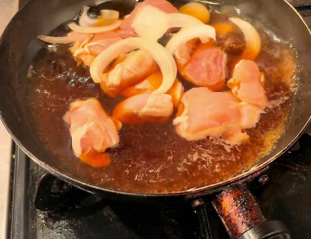 Morceaux de poulet et oignons émincés mijotant dans un bouillon sombre à la sauce soja dans une poêle noire.
