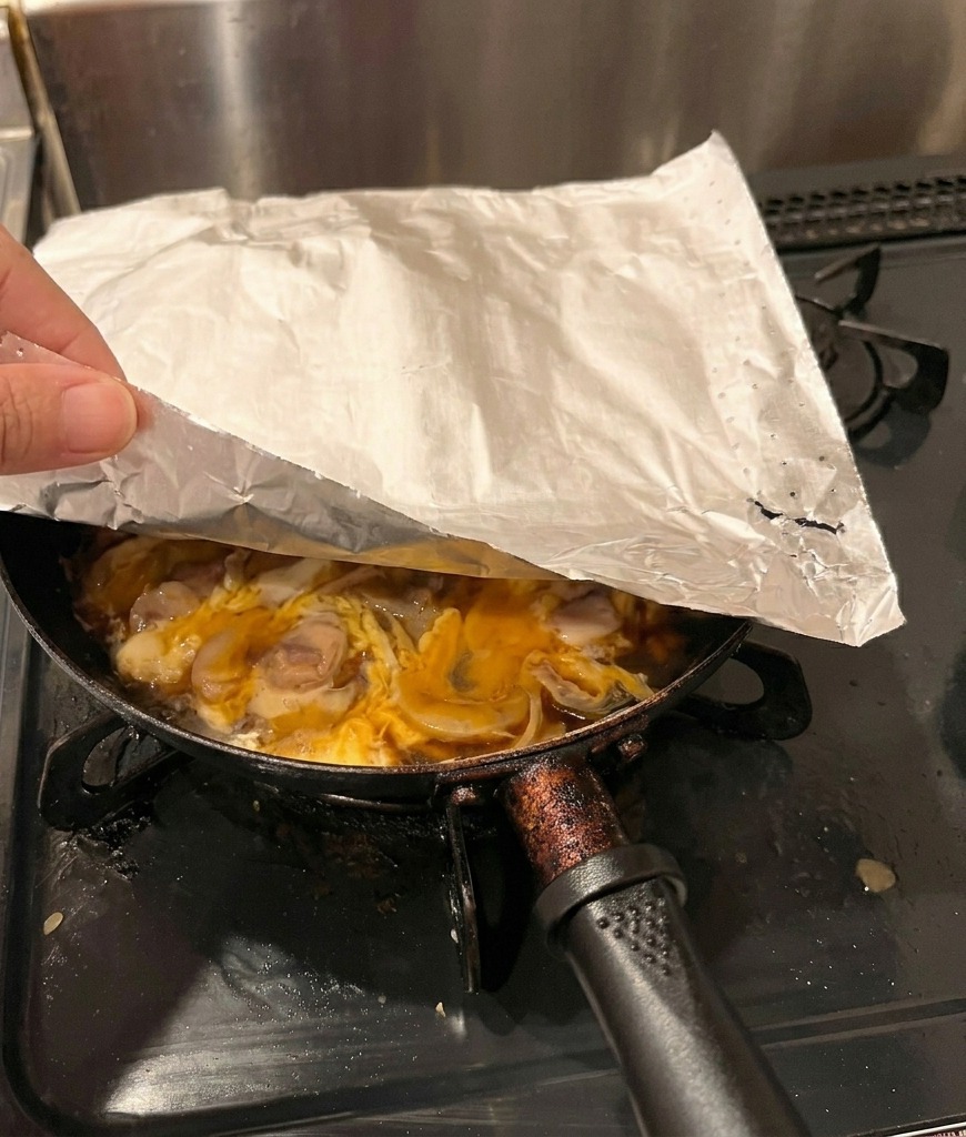 Une main plaçant une feuille de papier sulfurisé blanc sur une poêle contenant du poulet et des œufs partiellement cuits.
