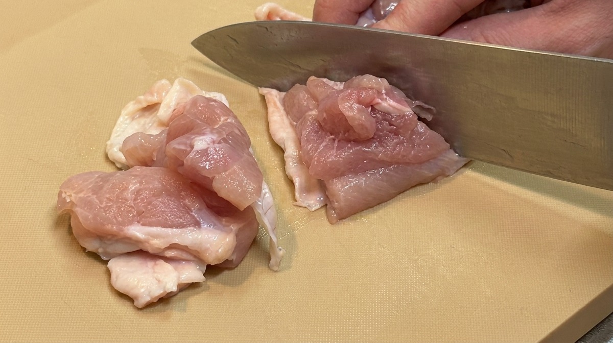 Des morceaux de hauts de cuisse de poulet crus coupés en dés réguliers avec un grand couteau de cuisine.