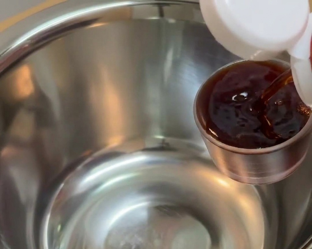 De la sauce soja foncée versée depuis un petit récipient doseur en métal dans un grand bol à mélanger en inox.