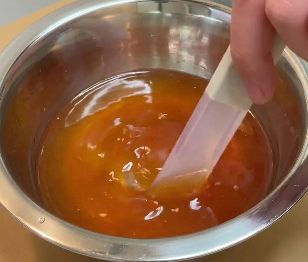 Une main utilisant une spatule pour mélanger un liquide dassaisonnement foncé et translucide dans un bol en inox.
