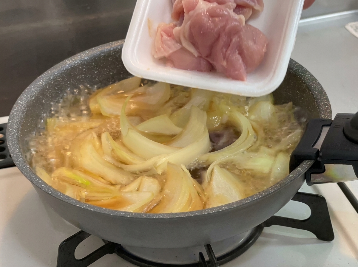 Des morceaux de hauts de cuisse de poulet crus et roses ajoutés depuis un plateau en polystyrène blanc dans une poêle contenant des oignons qui mijotent.