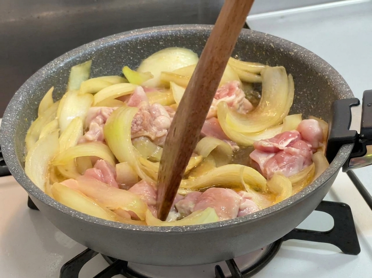 Une spatule en bois mélangeant des morceaux de poulet partiellement cuits et des oignons dans un liquide foncé frémissant.