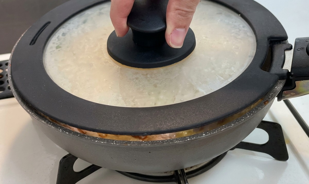 Une main plaçant un couvercle en verre doté dun bouton noir sur une poêle contenant du poulet, des oignons et des œufs.
