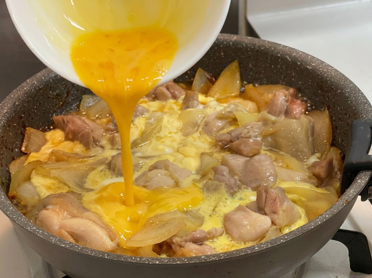 Des œufs battus dun jaune éclatant versés depuis un bol blanc dans une poêle chaude remplie de morceaux de poulet frémissants, doignons et dœufs partiellement figés.
