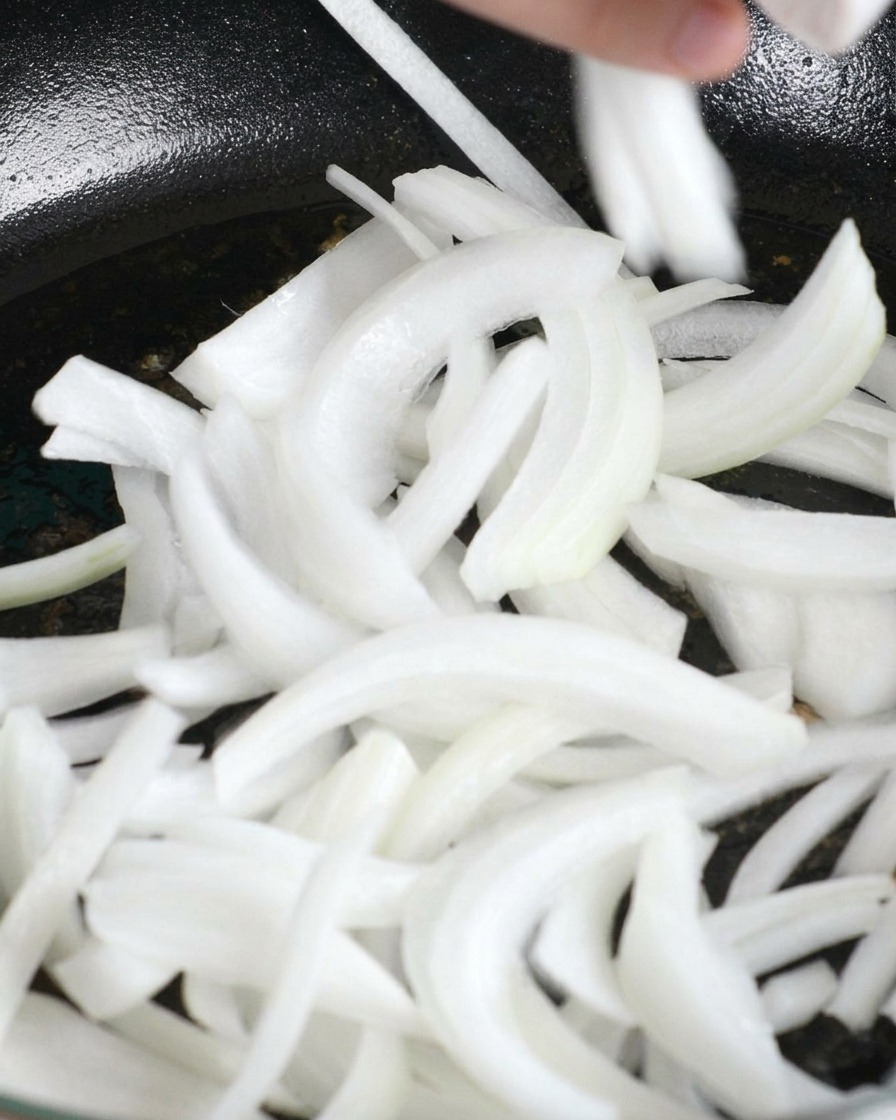 Des oignons blancs émincés ajoutés dans une poêle contenant le gras de cuisson du poulet.
