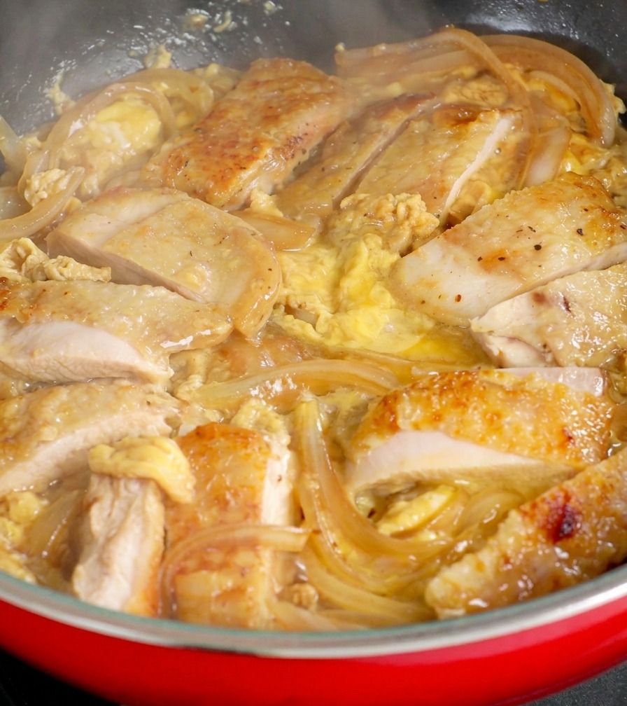 Des morceaux de poulet nichés parmi des oignons translucides dans une poêle, avec le reste doeuf cru versé par-dessus.