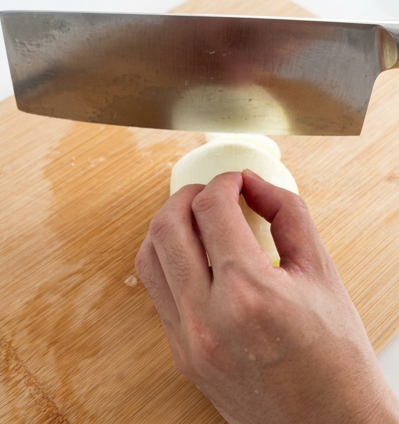Une main utilisant un grand couperet en acier inoxydable pour émincer un oignon blanc en fines lamelles sur une planche en bambou.