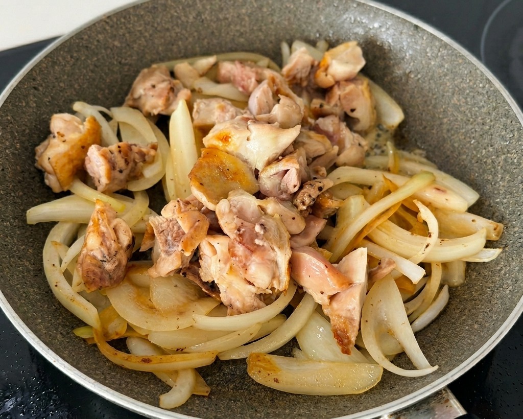 Lamelles doignons sautées mélangées à des morceaux de poulet dorés et des oignons verts émincés dans une poêle antiadhésive.