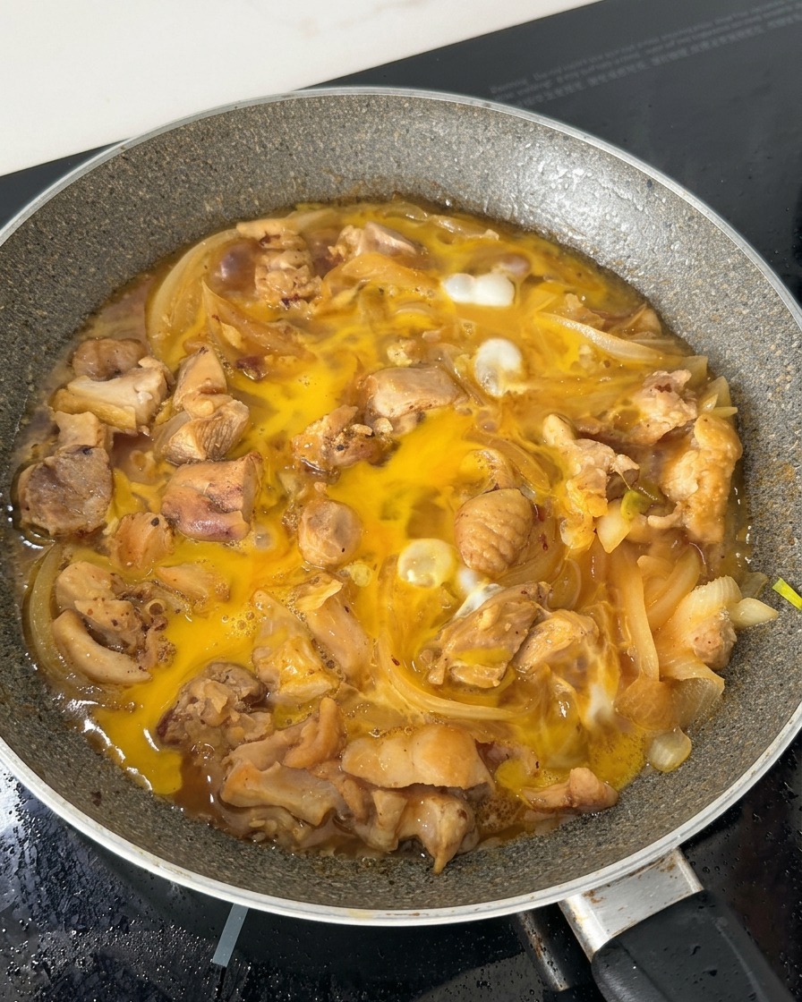 Mélange dœufs partiellement cuits bouillonnant dans une poêle avec des morceaux de poulet et des oignons émincés dans un bouillon de soja.