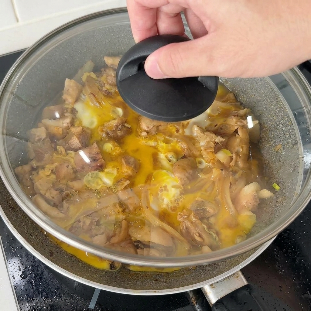 Une main plaçant un couvercle en verre sur une poêle de poulet et dœufs pour cuire à létouffée le reste du mélange dœufs.