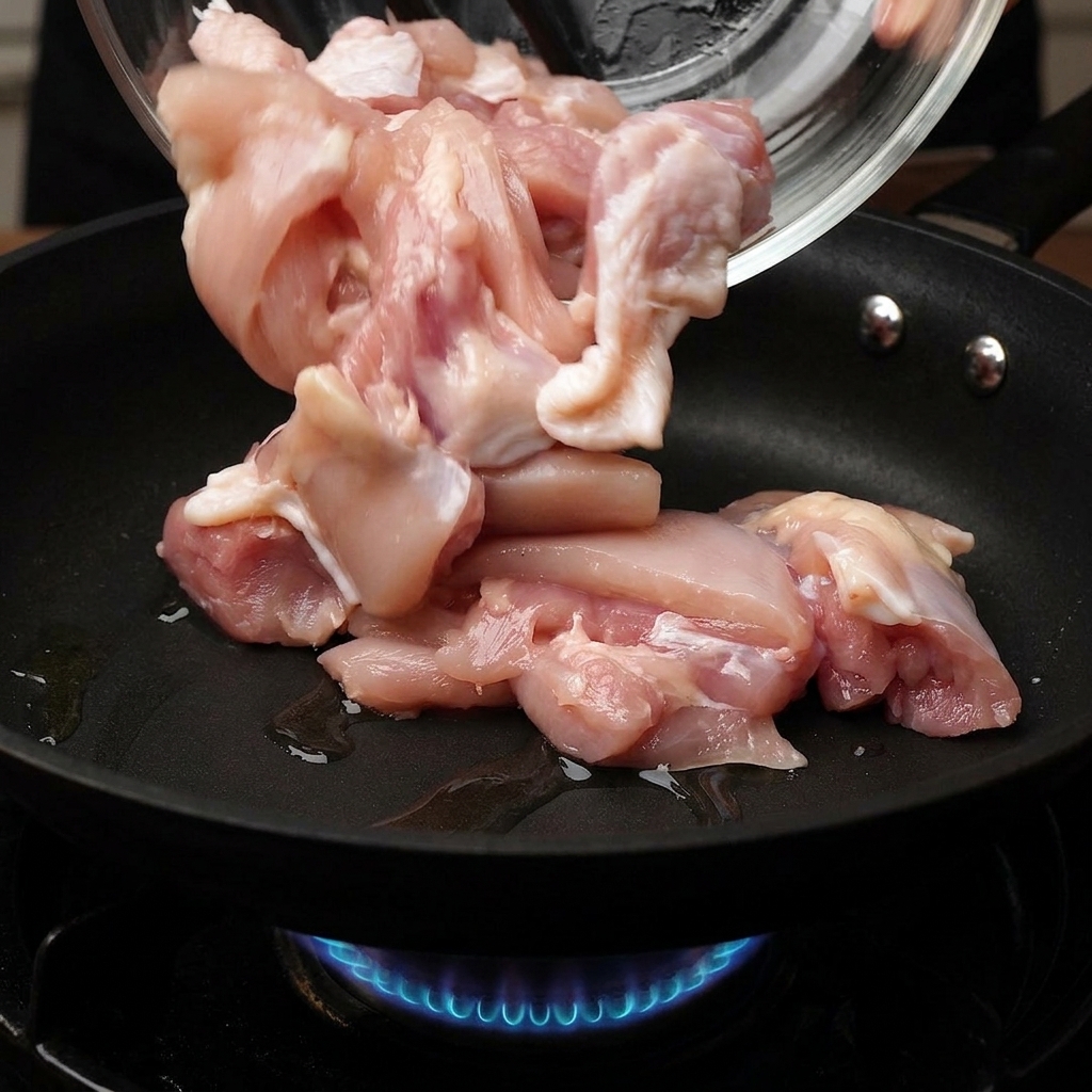 Morceaux de poulet cru versés dun bol en verre dans une poêle chaude, avec une flamme de gaz bleue visible en dessous.