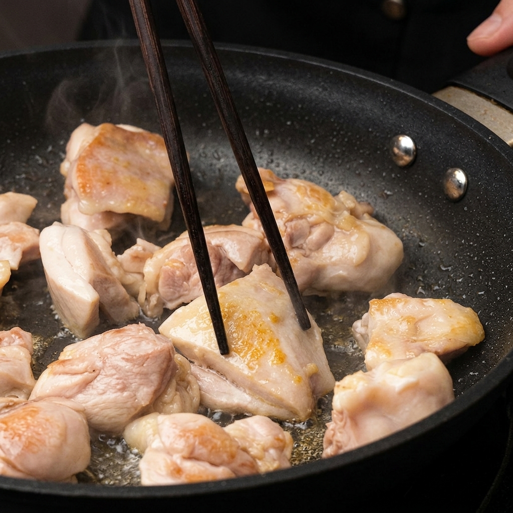 Morceaux de poulet dorant dans une poêle, retournés à laide de baguettes en bois sombre.