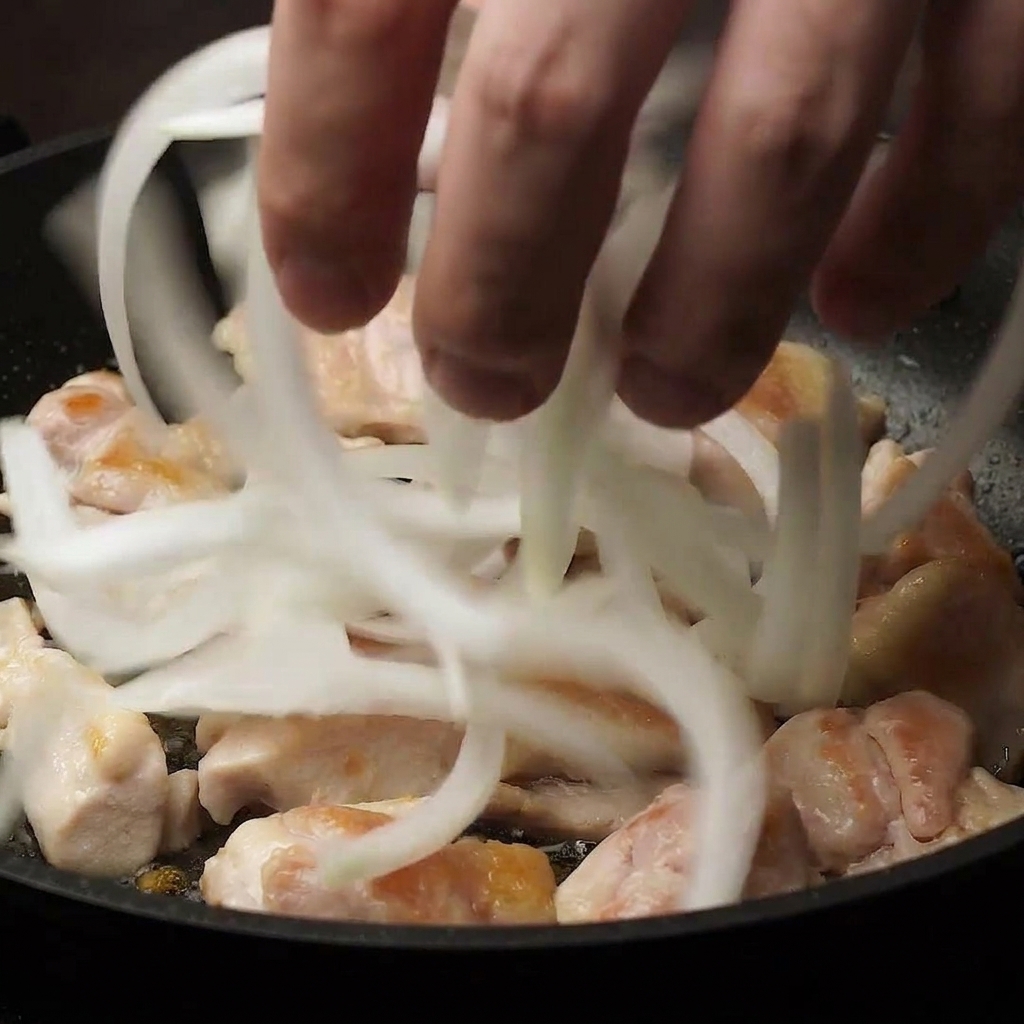 Une main ajoutant des oignons émincés dans une poêle contenant les morceaux de poulet mi-cuits.
