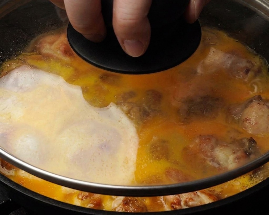 Gros plan dune poêle avec un couvercle en verre montrant les œufs battus recouvrant partiellement les morceaux de poulet en cours de cuisson.