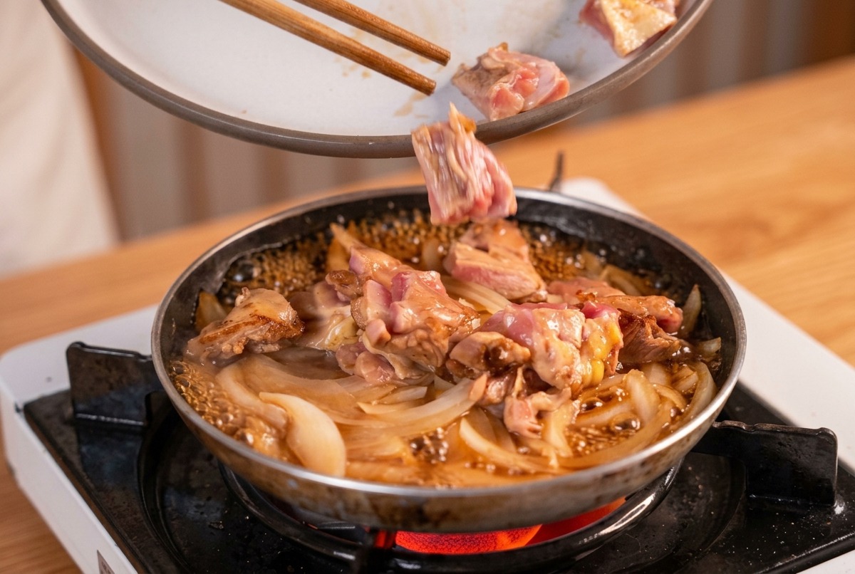 Morceaux de poulet saisis ajoutés avec des baguettes dans une poêle doignons et de bouillon dashi frémissant.