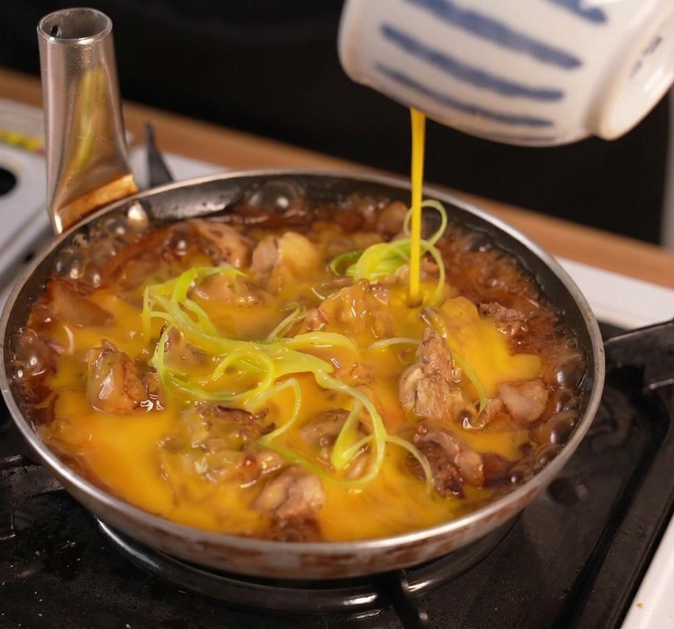 Œuf battu versé dun bol à rayures bleues et blanches sur du poulet et des oignons verts mijant dans une sauce foncée.