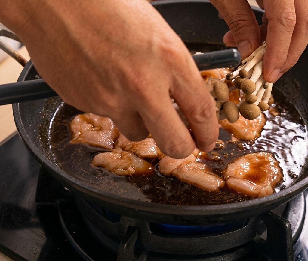 Des mains déposant une poignée de champignons shimeji dans une poêle de poulet qui mijote dans la sauce sombre.